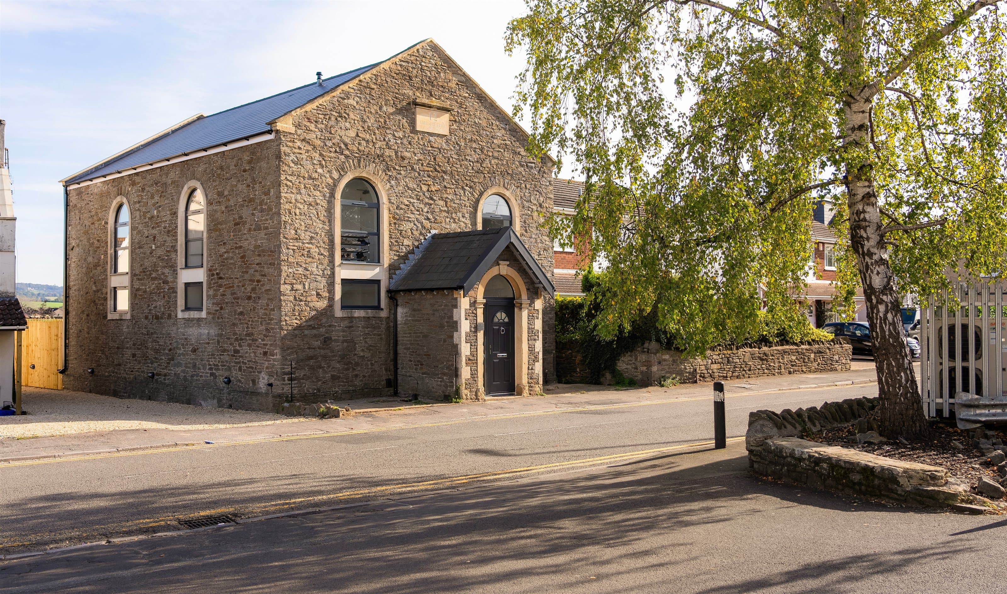 Converted stone chapel with arched windows and a gabled entrance porch, beside a tree-lined street